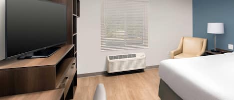 Une chambre d’hôtel avec un lit, un téléviseur posé sur un meuble en bois, une chaise, un bureau, une lampe et une fenêtre avec des stores.