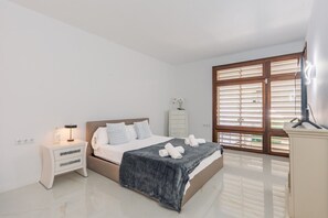 Een slaapkamer met een bed, nachtkastje, kaptafel en een groot raam met zonwering.