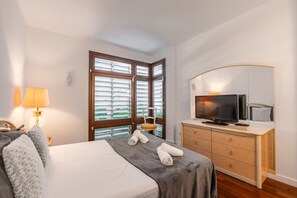 A hotel room with a bed, a television on a wooden cabinet, a window with blinds, and a lamp on the left. - Villa Bella - Exclusive Roses Retreat (Girona)
