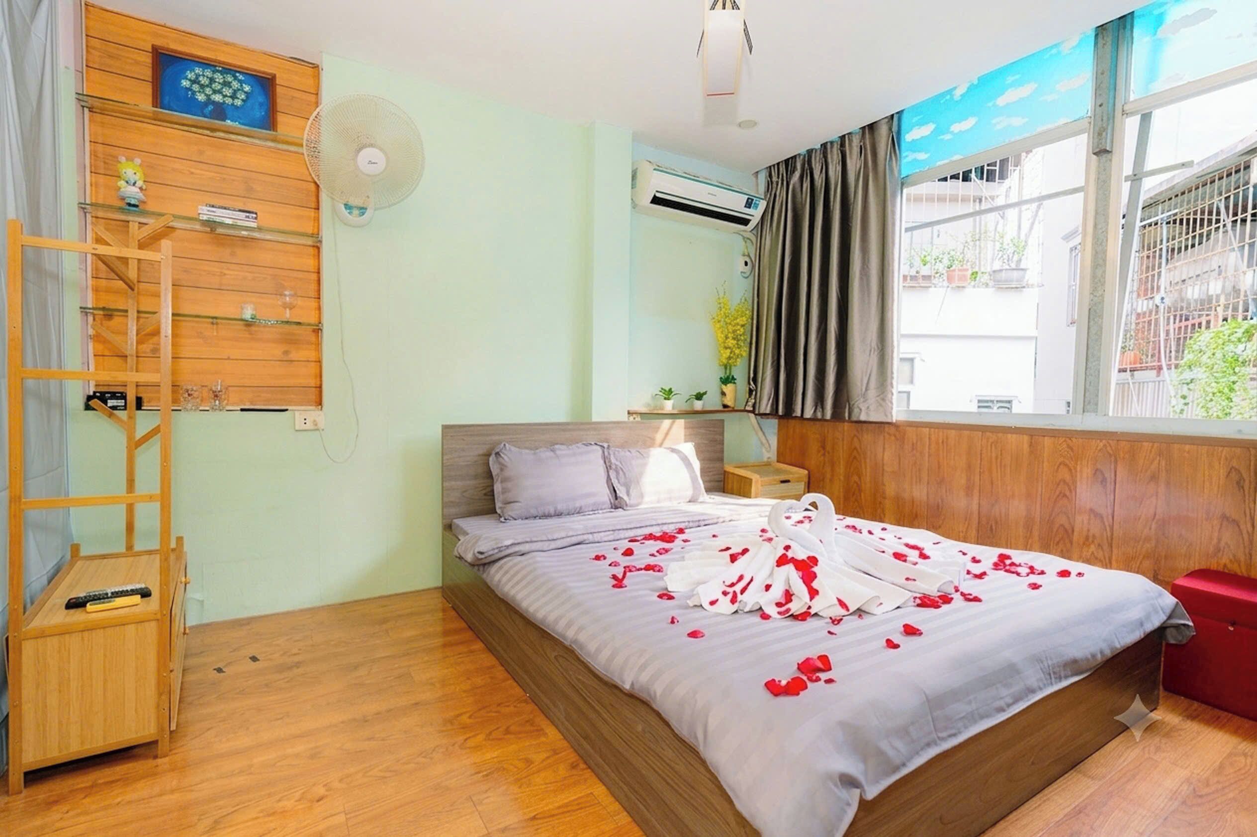 A bedroom with a bed, wooden floor, a fan, an air conditioning unit, a small shelf with items, and a window with curtains.
