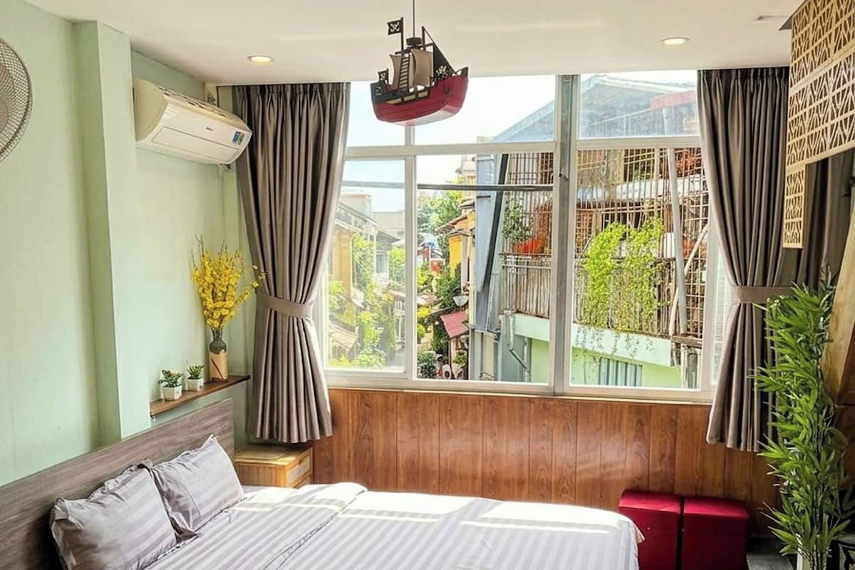 A bedroom with a large bed, wooden flooring, a window with curtains, and a view of buildings outside.