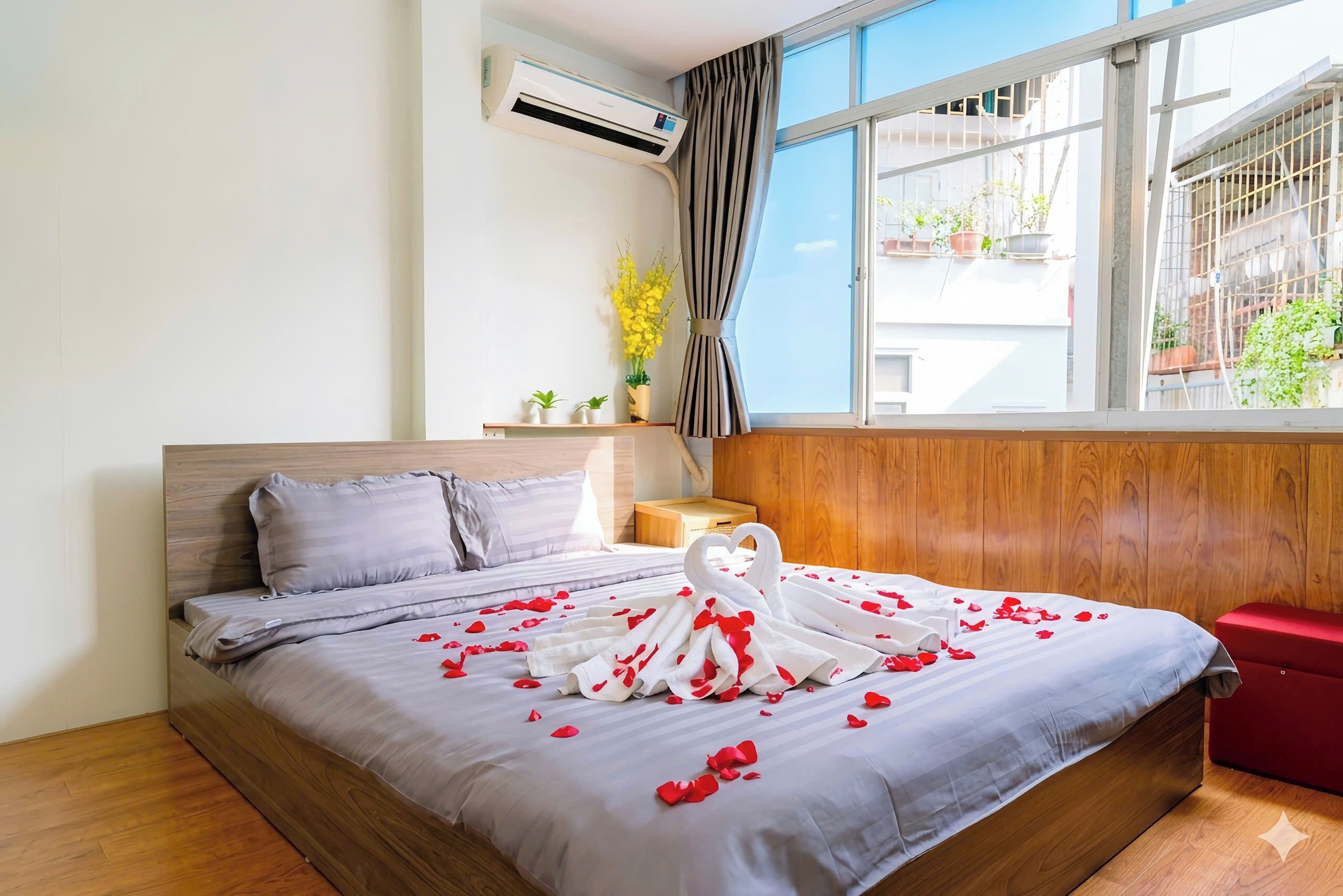 Habitación de hotel con cama, cabecera de madera, mesita de noche, silla roja, ventana con cortinas y una planta en maceta.