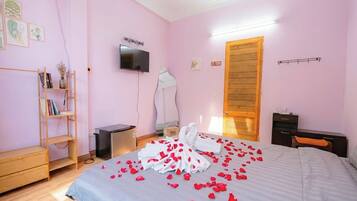 A bedroom with a bed, a TV, a wooden wardrobe, a shelf with books and a vase, and a door with a towel rack.