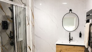 Un baño moderno con cabina de ducha de vidrio, pared de mármol, tocador de madera y espejo redondo.