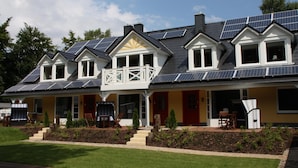 Uma casa de dois andares com painéis solares, uma varanda e um jardim bem cuidado.