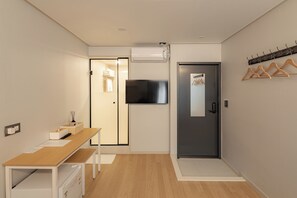 A hotel room with a wooden desk, a flat-screen TV mounted on the wall, a door with a coat rack, and a small table with a tissue box and remote control.