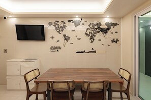 Uma sala de jantar com mesa de madeira e quatro cadeiras, um mapa-múndi na parede, televisão montada acima da mesa e um separador de vidro à direita.
