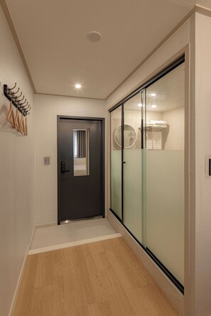 Um corredor com uma porta escura, um cabideiro com cabides de madeira e uma porta de vidro deslizante que dá para um banheiro com um porta-toalhas.
