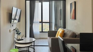 A modern living room with a sofa, a TV mounted on the wall, a small table, and a window with curtains. - The Rich Rama 9 - Srinakarin (Khet Suan Luang)