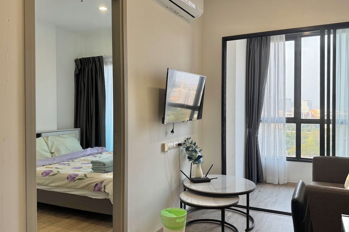 A hotel room with a bed, a TV mounted on the wall, a small table, a chair, and a window with curtains.