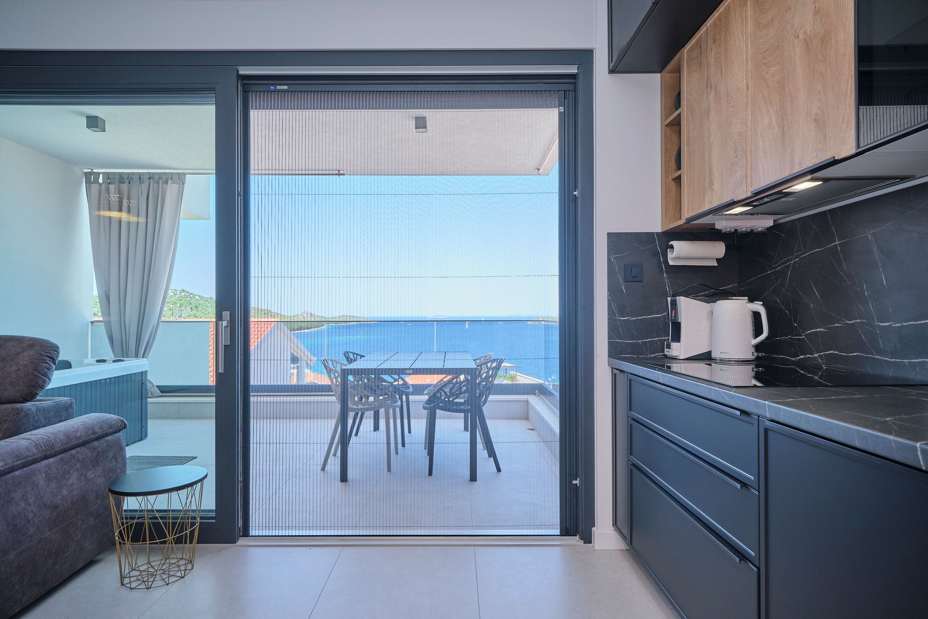 A modern kitchen with a black marble countertop, a coffee machine, and a view of the sea through a sliding glass door.
