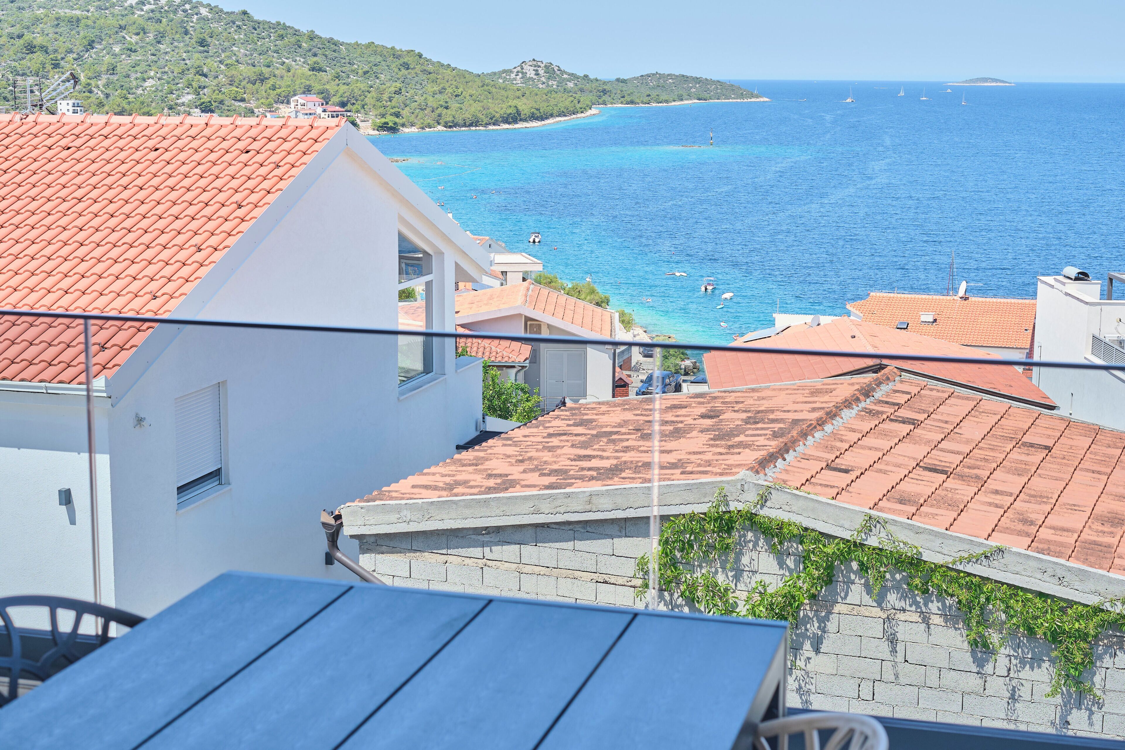 A coastal view with terracotta roofs, a clear blue sea, and a hilly landscape in the distance.