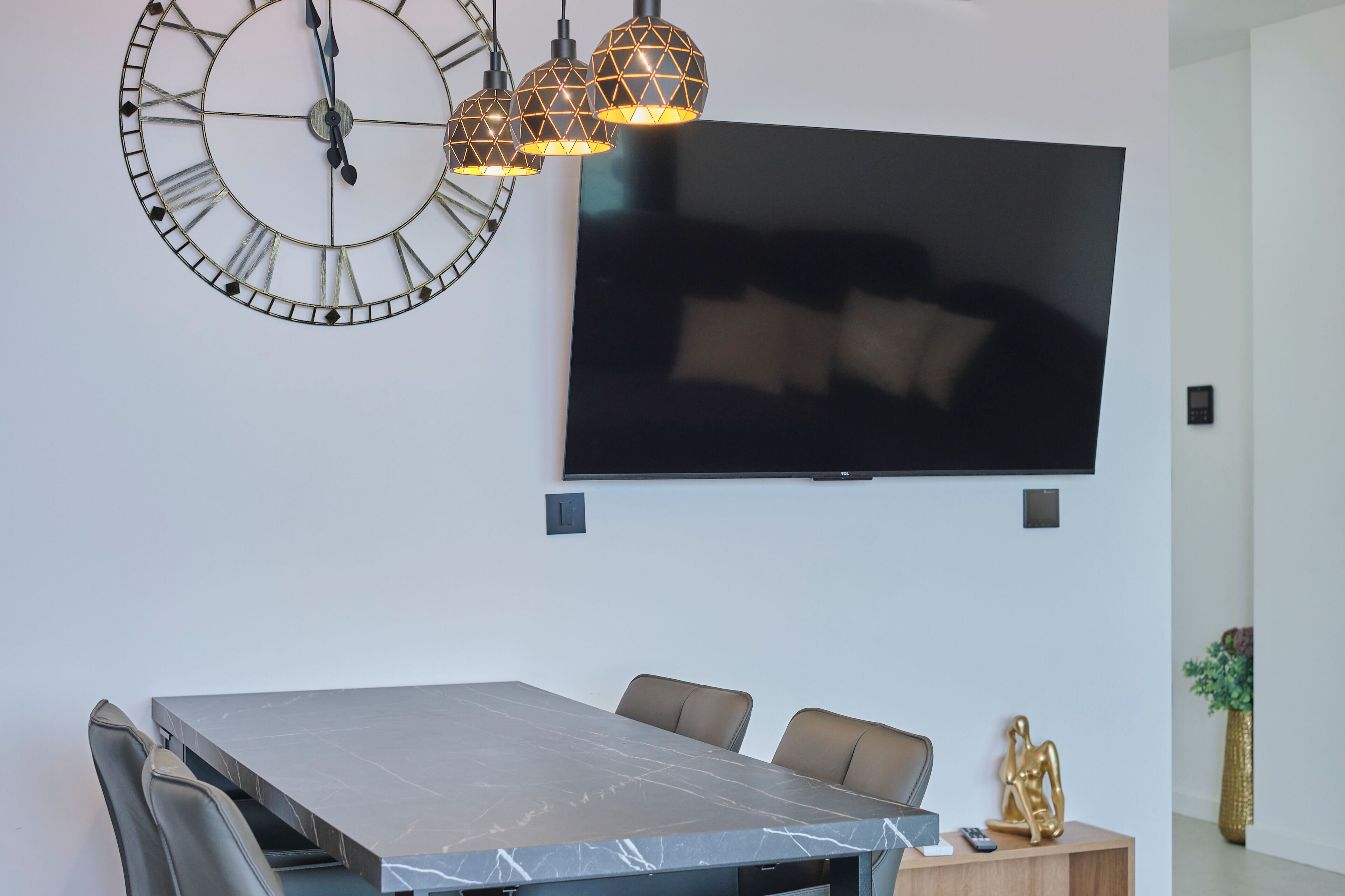 A modern dining area with a marble table, leather chairs, a large flat-screen TV, and a decorative wall clock.