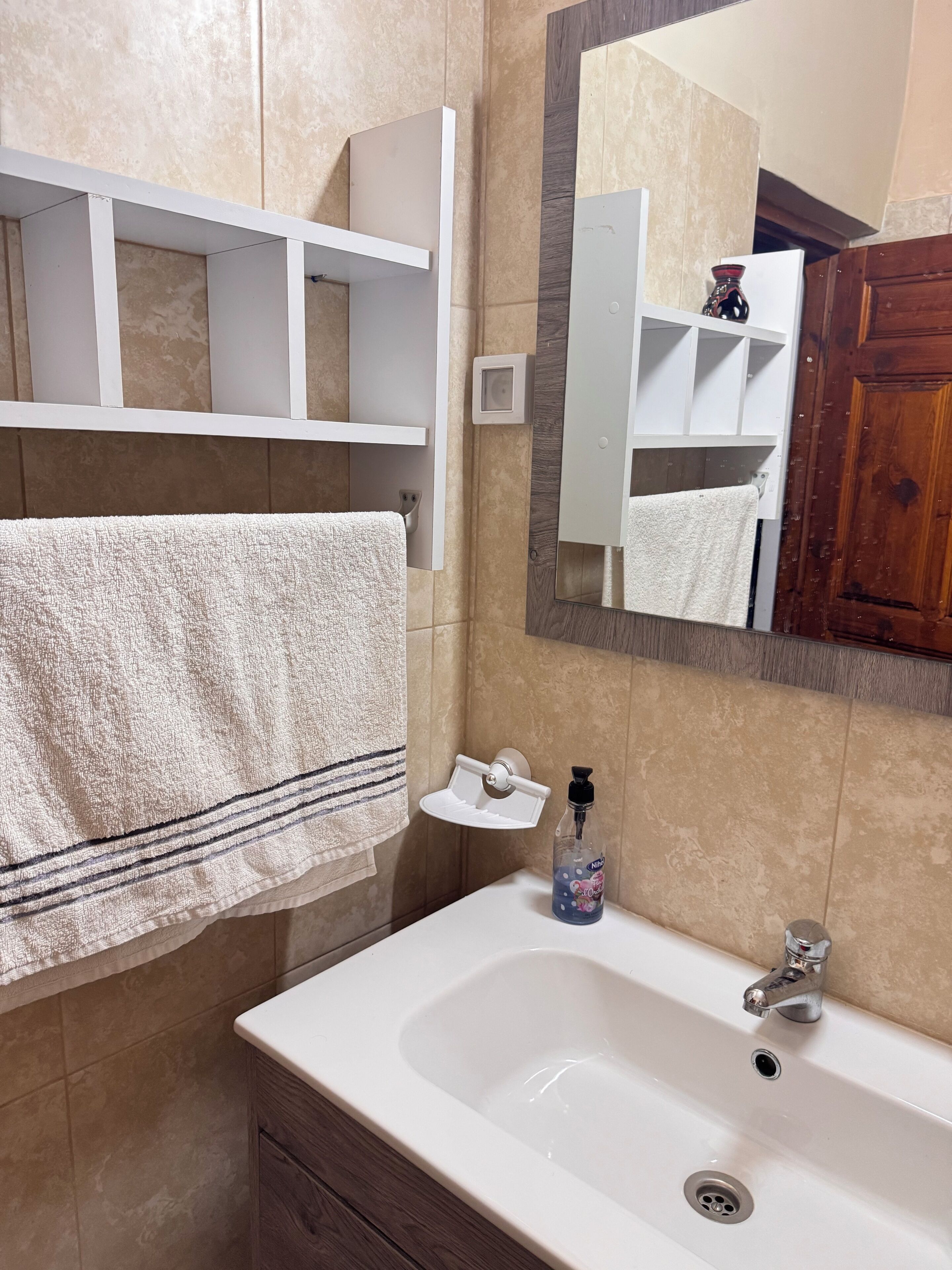A bathroom with a white sink, a mirror, a towel hanging on the wall, and a shelf above the sink.
