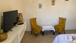 A room with a bed, a television on a stand, two chairs, a small table, and a decorative vase.