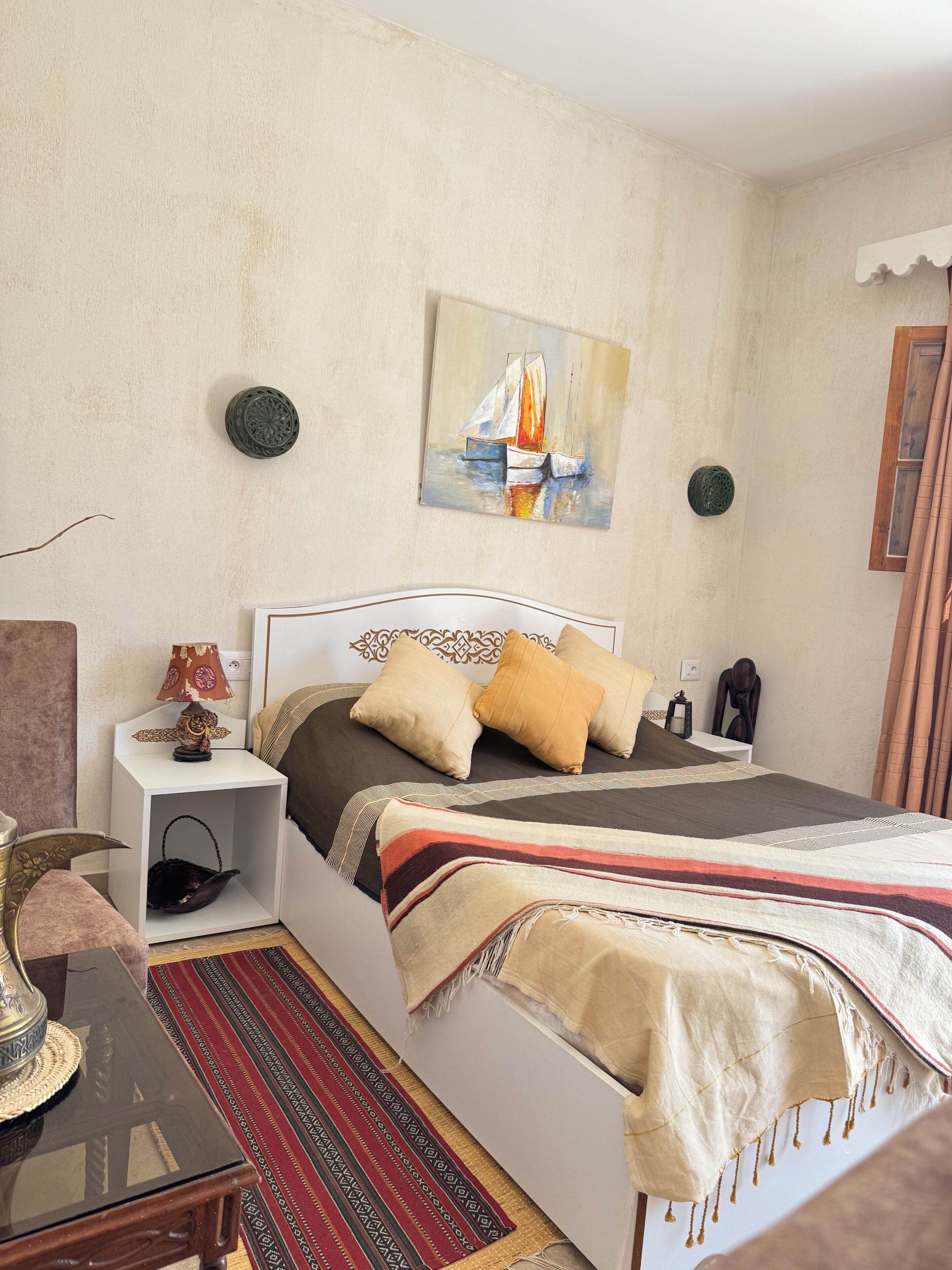 Quarto com cama, mesa de cabeceira, luminária, almofadas decorativas, um quadro e um tapete estampado.