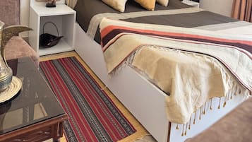Quarto com cama, mesa de cabeceira, luminária, almofadas decorativas, um quadro e um tapete estampado.