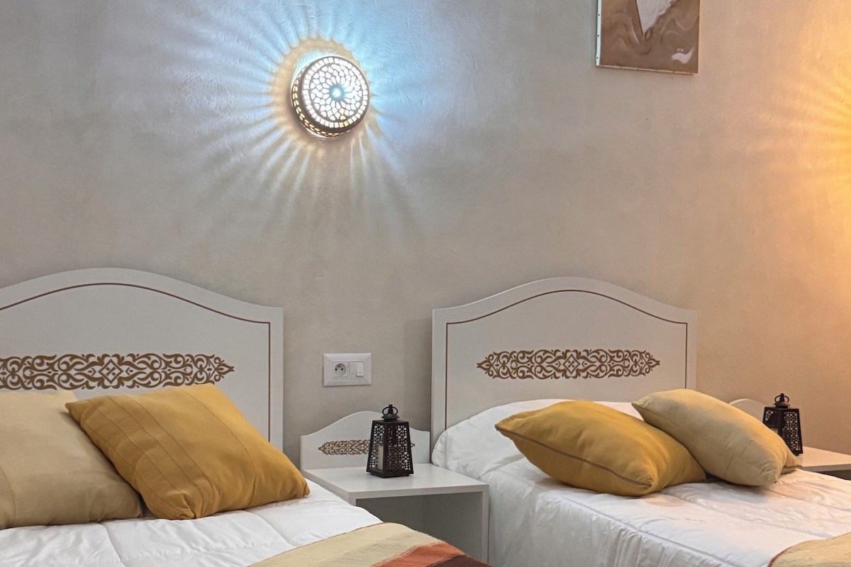 Une chambre d’hôtel avec deux lits, une table de chevet, un luminaire fixé au mur et un tableau encadré.