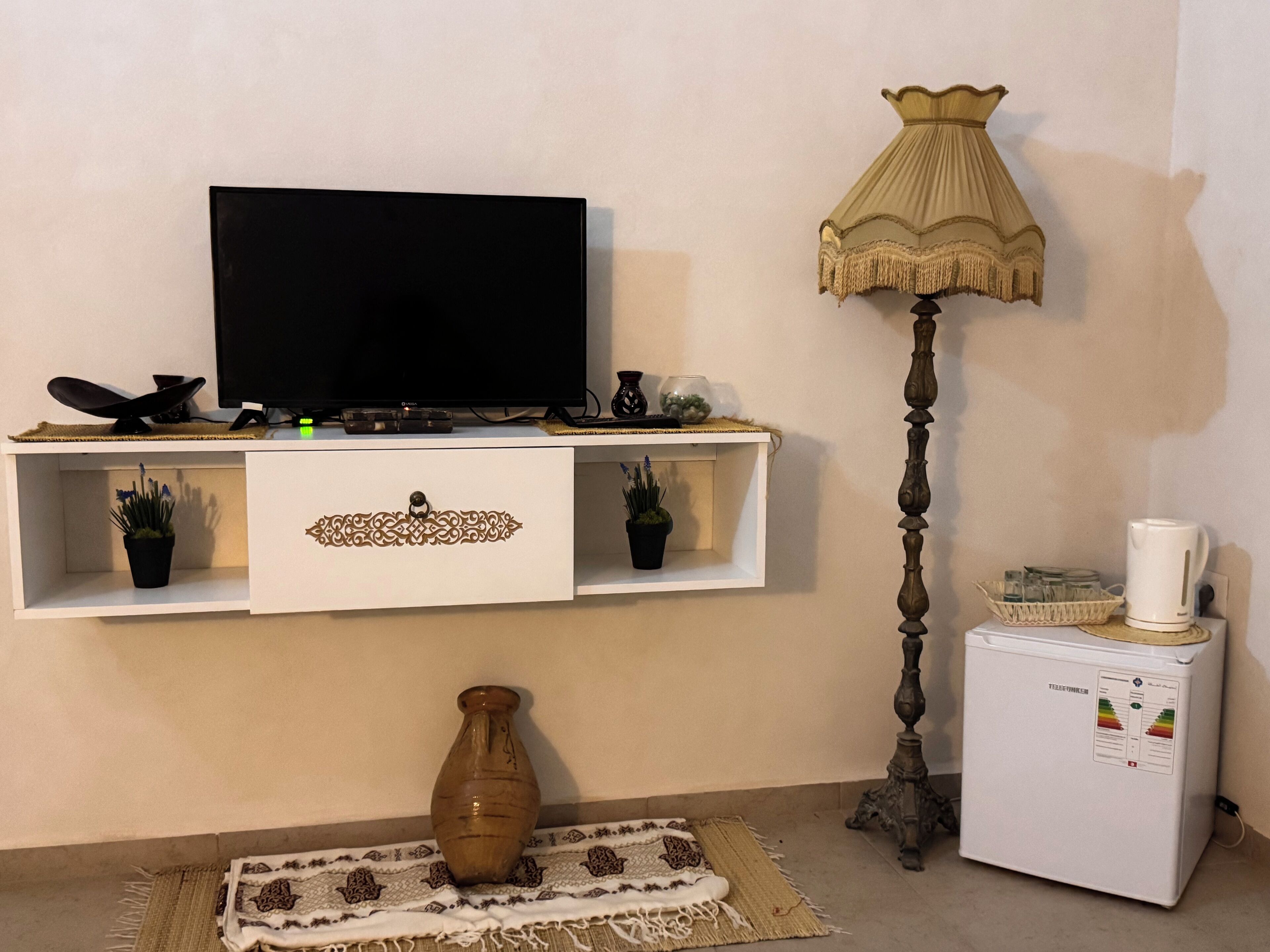 A living room with a flat-screen TV, a lamp, a coffee maker, a vase, and a small refrigerator.