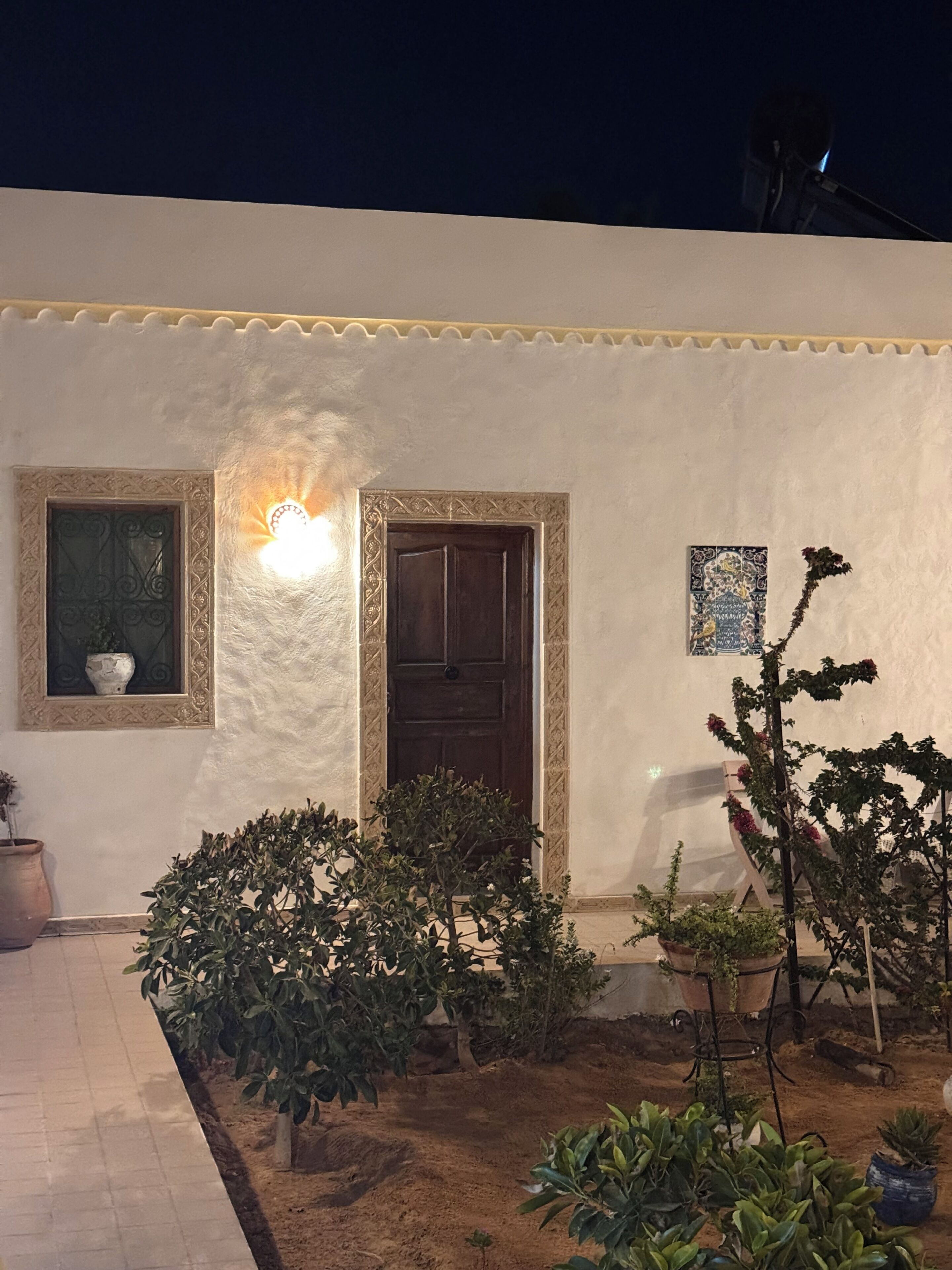 Un edificio blanco con una puerta y ventanas, rodeado de plantas en macetas y un pequeño jardín.