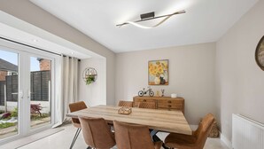 A dining area with a wooden table, brown chairs, a radiator, and a painting of sunflowers. - Spacious Modern home in Whitefield - Sleeps 9 (Whitefield)