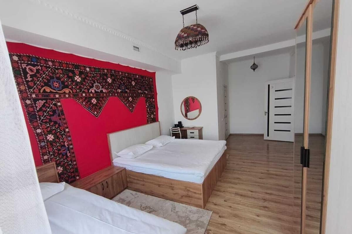 Quarto com duas camas, tapeçaria vermelha, piso de madeira, espelho e guarda-roupa.