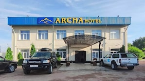 Un hotel con fachada azul y blanca, un acceso cubierto y dos vehículos estacionados al frente.
