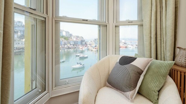 Una stanza con ampie finestre, vista su un porto con barche e un angolo salotto accogliente con cuscini.