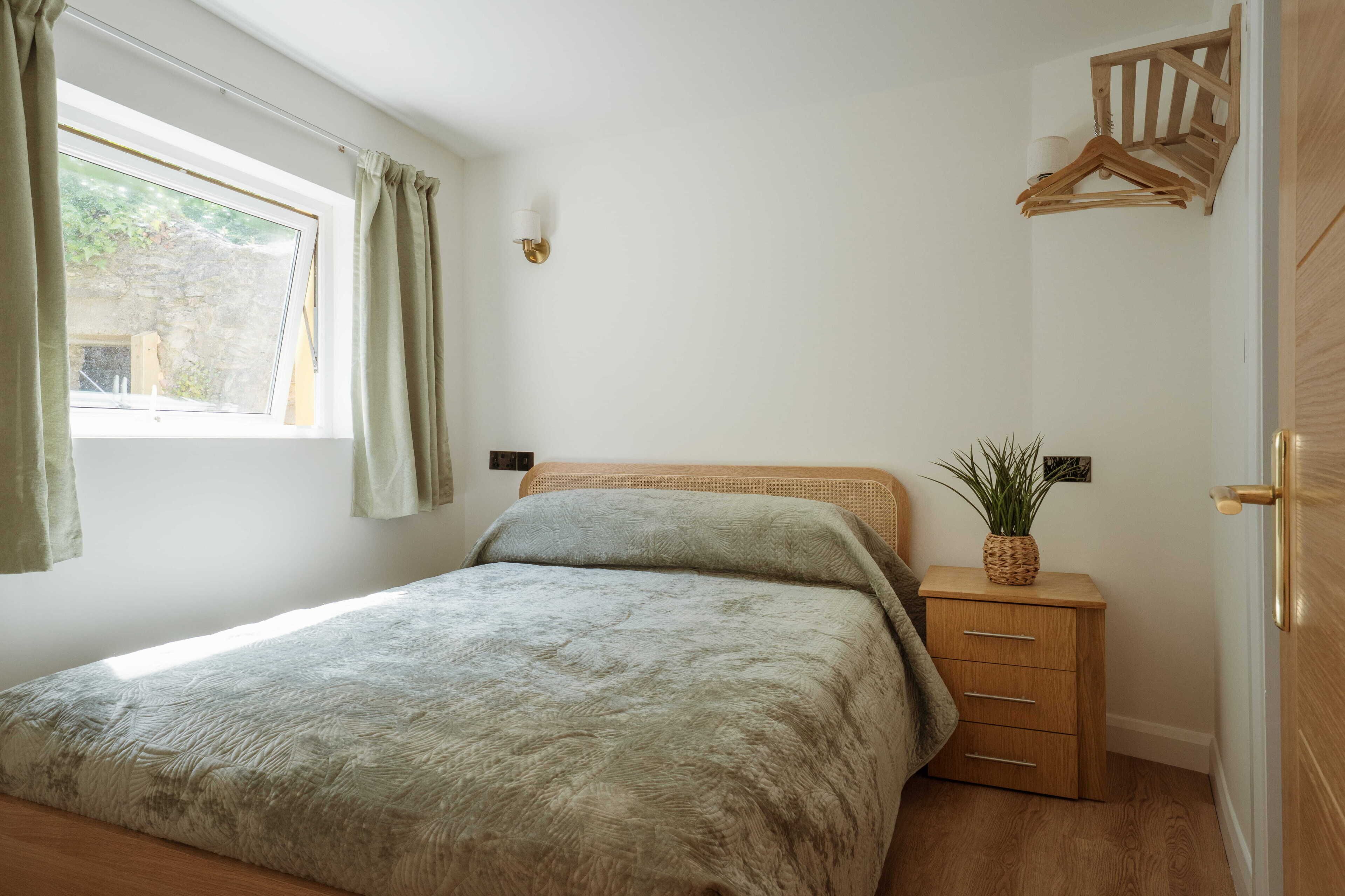 A bedroom with a bed, a bedside table with a plant, a window with curtains, and a wooden door.