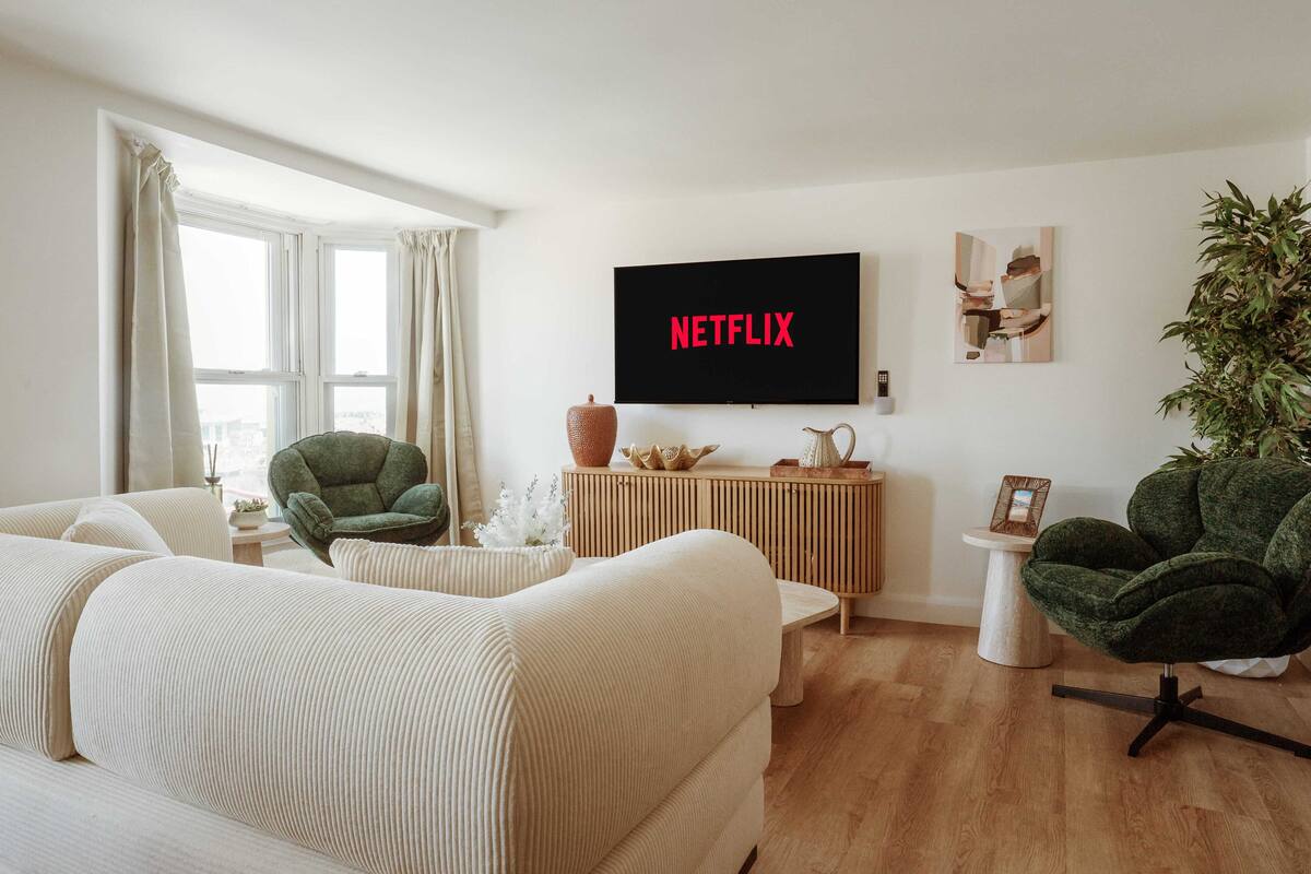 Un salón con sofá, sillones, un televisor que muestra 'NETFLIX', un aparador de madera y una planta en maceta.