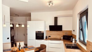 A compact kitchen with a dining area, wooden flooring, a built-in oven, a black TV mounted on the wall, and a window with a view. - Serenity House—Private Infinity Pool, Panoramic Sea Views, and Near Budva (Krimovica)