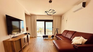 A modern living room with a flat-screen TV, wooden flooring, a brown leather sofa, and a balcony with a view. - Serenity House—Private Infinity Pool, Panoramic Sea Views, and Near Budva (Krimovica)
