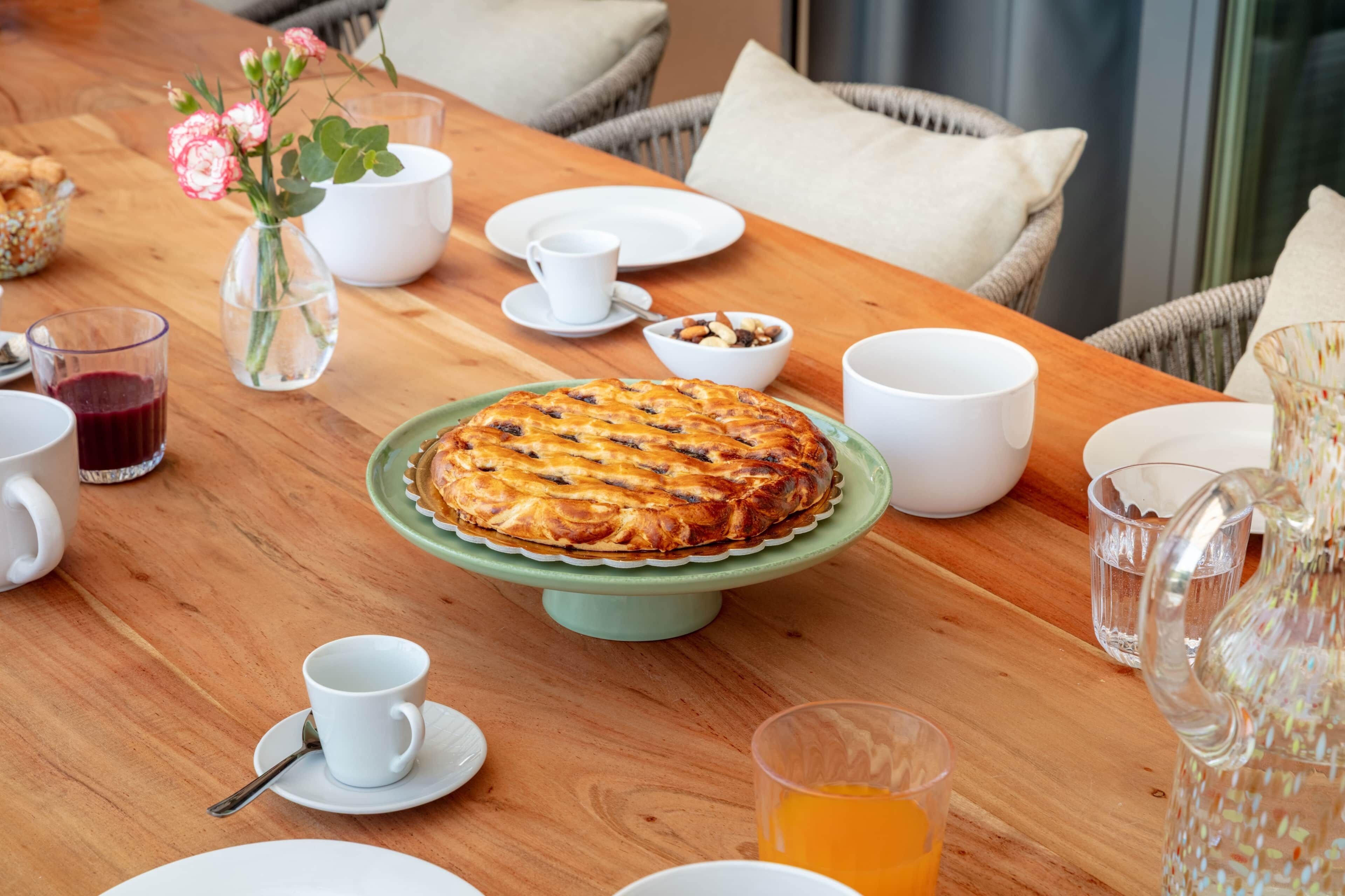 Een houten eettafel met een taart op een groene standaard, witte borden, bekers, een schaal met noten, een vaas met bloemen en een karaf.