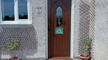 Uma porta marrom com uma placa de 'Welcome' e uma placa de 'The Nook', ladeada por duas plantas em vasos e um cercado treliçado.