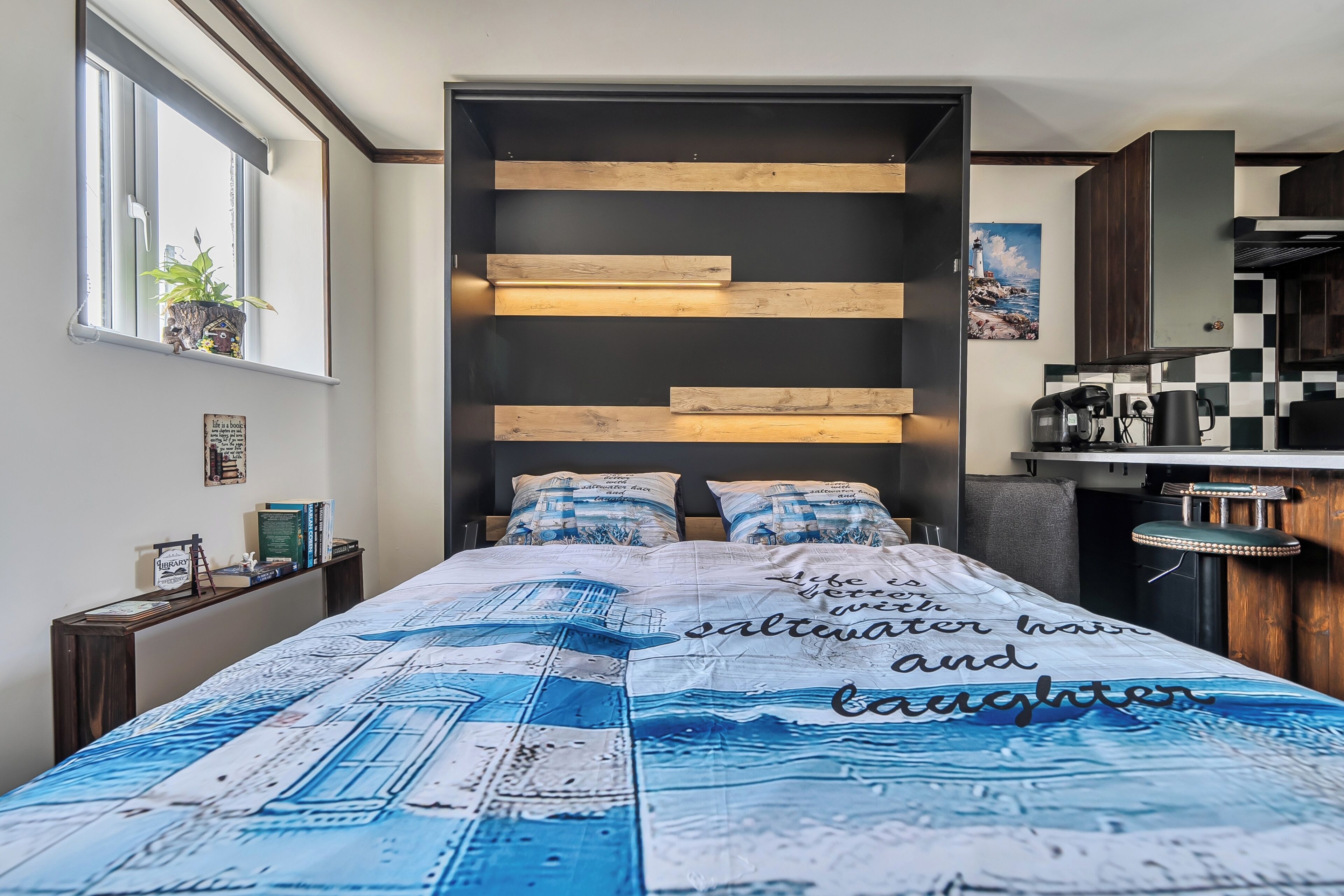 A modern bedroom with a bed featuring a nautical-themed bedspread, a built-in shelf with books, and a window with a plant.