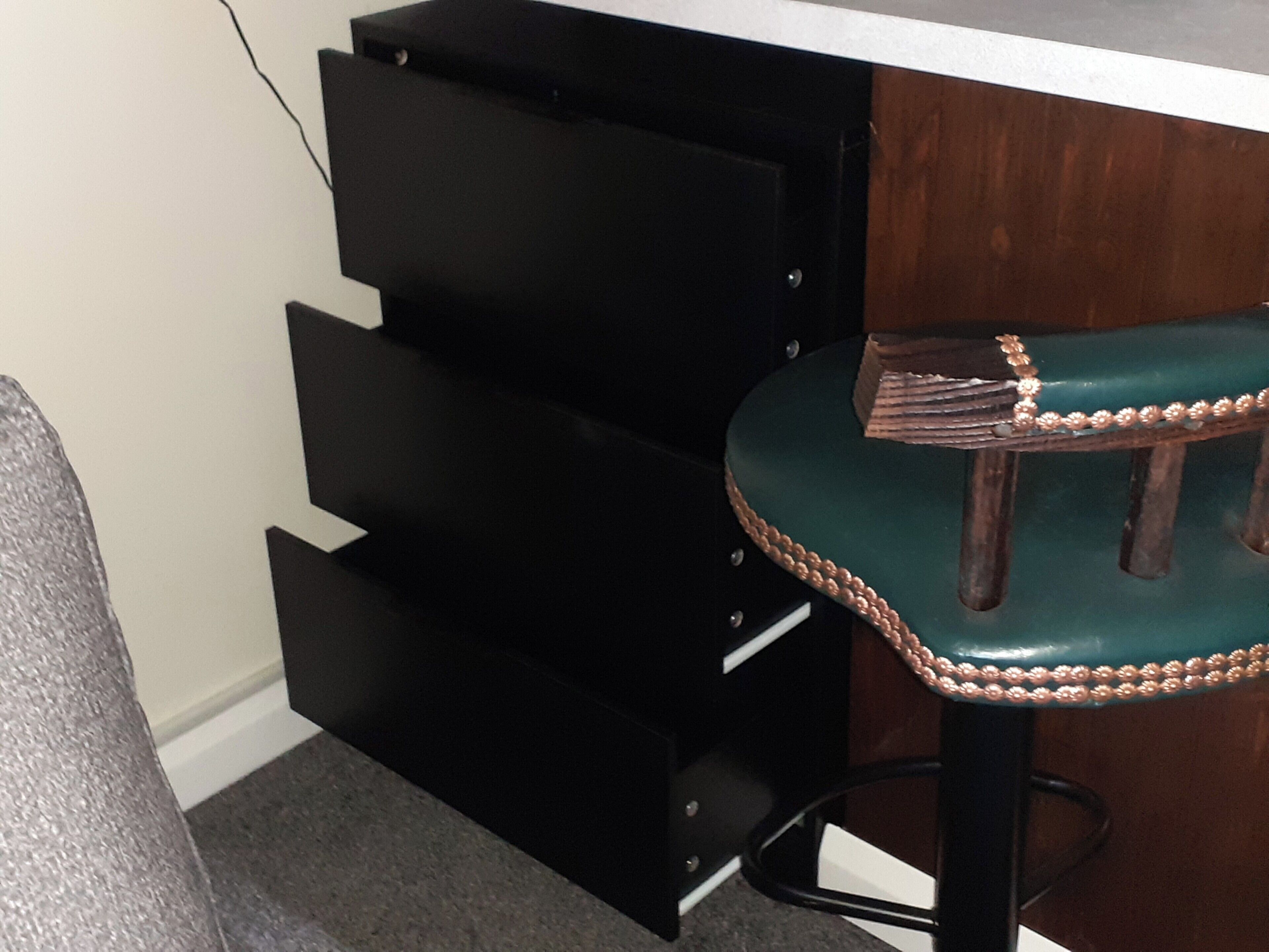Un tabouret de bar avec un siège vert et un bord décoratif, un comptoir de bar en métal noir doté d’un meuble à tiroirs en dessous, et un plan de travail blanc.