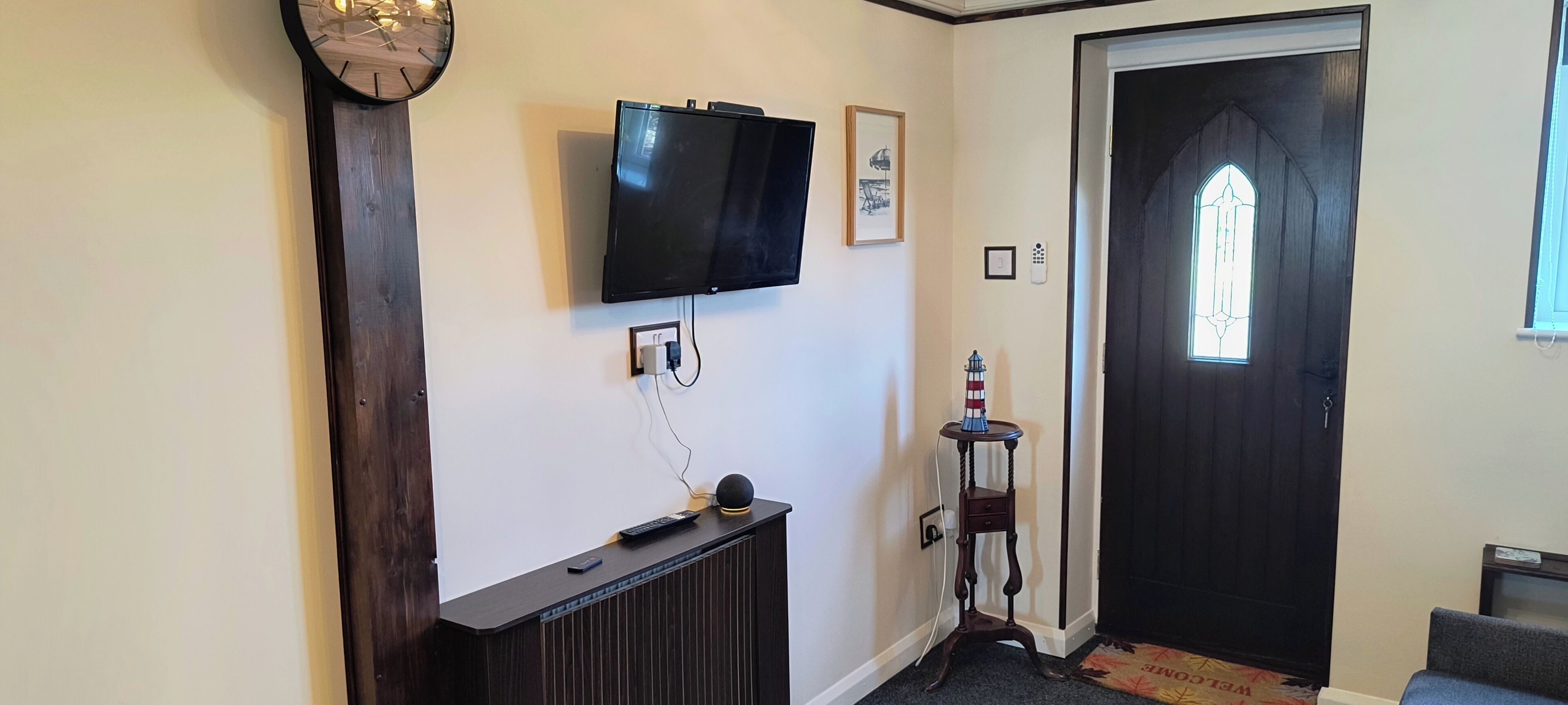 Un corridoio con una televisione a schermo piatto montata a parete, un tavolino consolle in legno, un piccolo tavolo con un modellino di faro e una porta scura con una finestra.