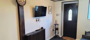 Un pasillo con televisor de pantalla plana montado en la pared, una consola de madera, una mesita con un modelo de faro y una puerta oscura con ventanal.