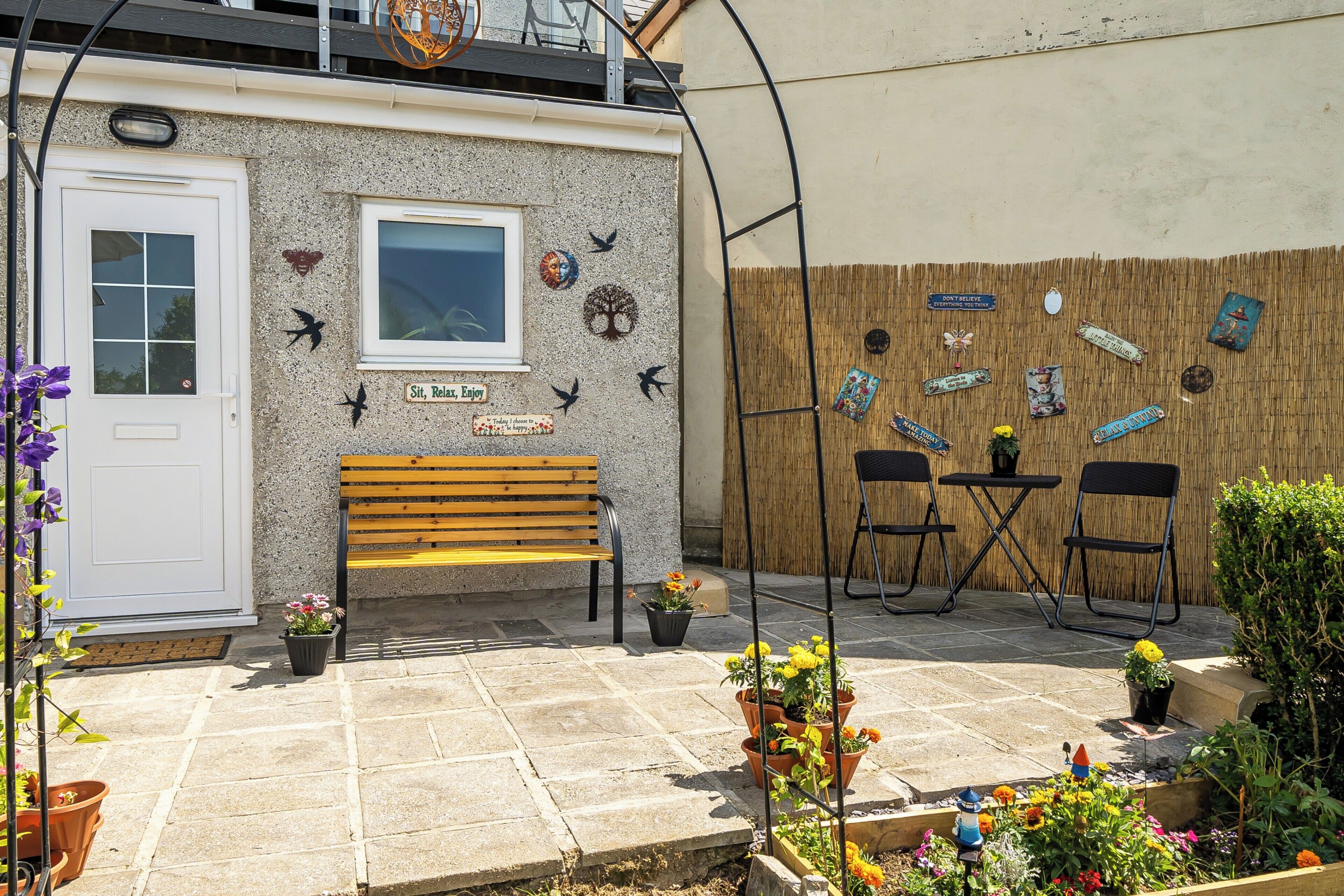 A small outdoor seating area with a bench and chairs, surrounded by potted plants and decorative wall art.