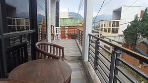 A balcony with a wooden table and chairs, a railing, and a view of buildings and mountains in the distance.