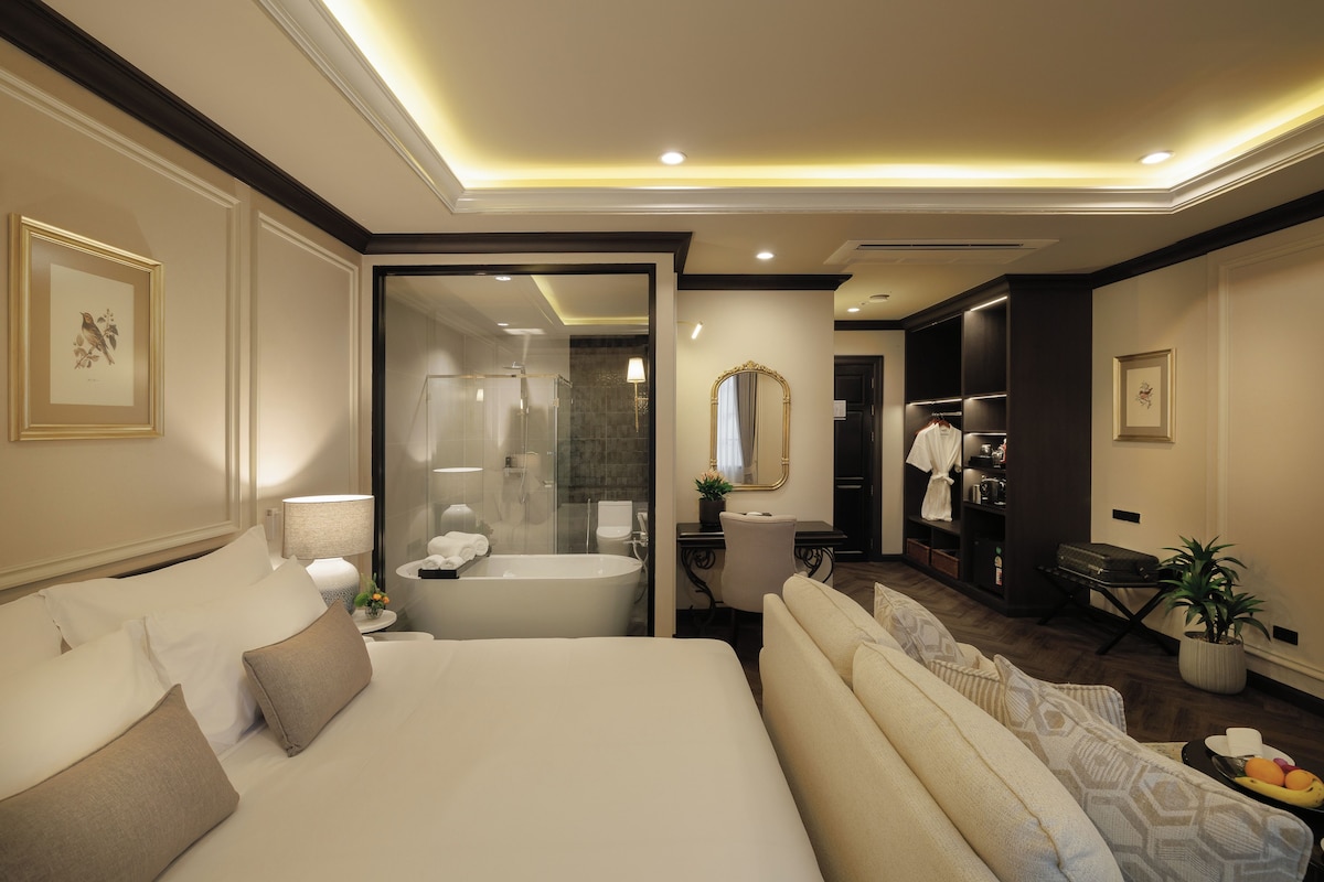 A modern hotel room with a large bed, a sitting area, a bathroom with a bathtub, and a walk-in closet.