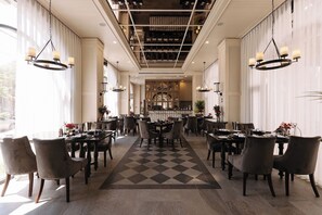 Un comedor de hotel con varias mesas y sillas, un tapete de piso a cuadros y candelabros colgantes.