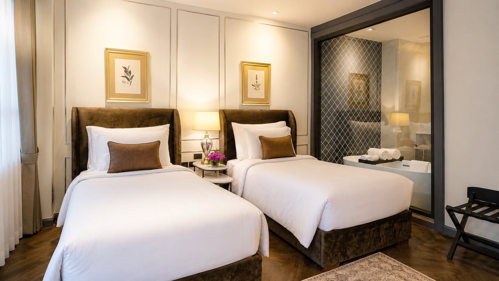 A hotel room with two beds, a nightstand with a lamp, a mirror, and a view of the bathroom.
