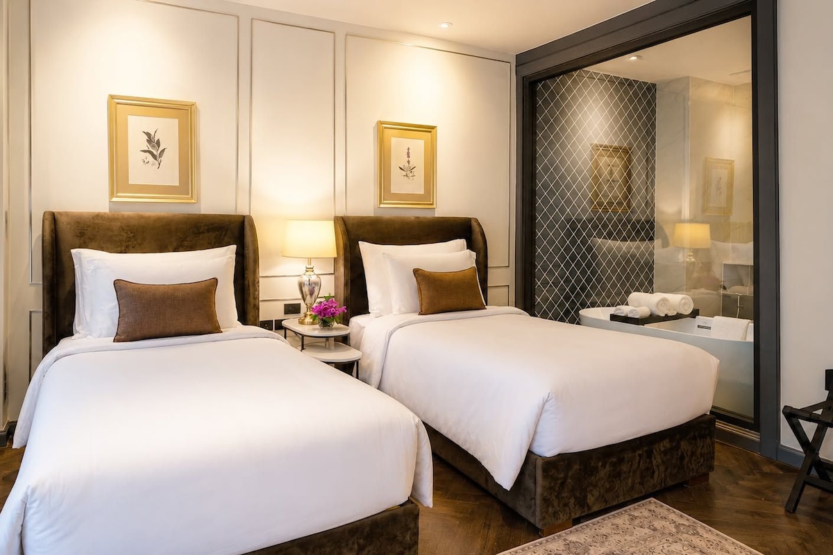 A hotel room with two beds, a nightstand with a lamp, a mirror, and a view of the bathroom.
