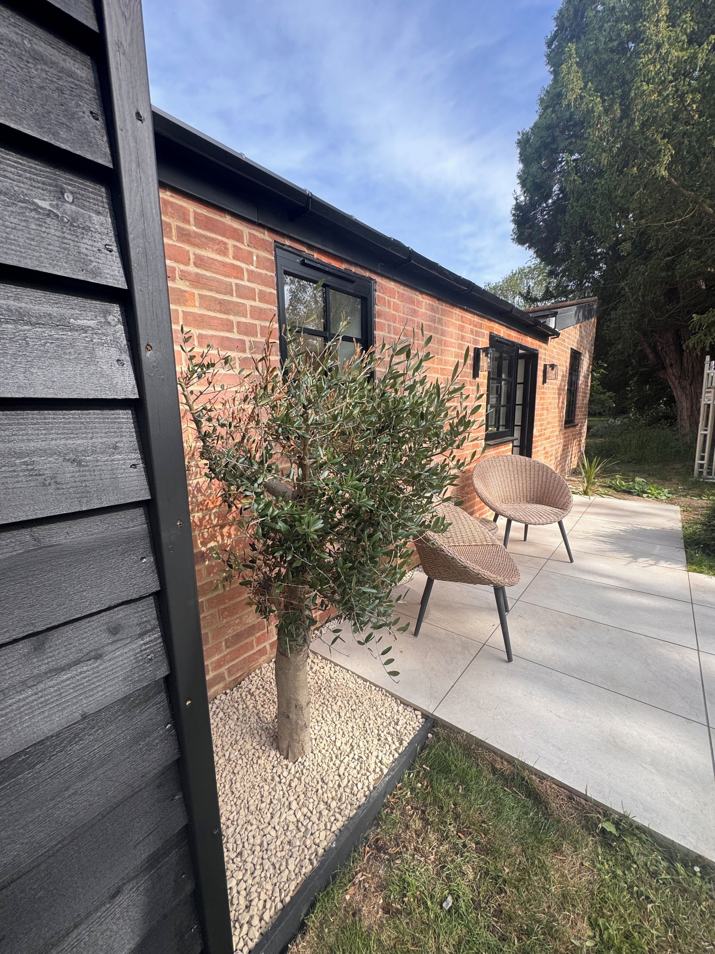 Un bâtiment en briques avec une fenêtre à cadre noir, un petit arbre et deux chaises en osier sur un patio en béton.