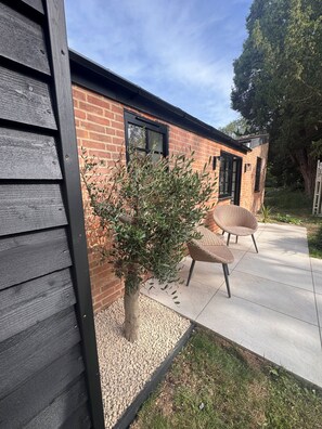 Un edificio de ladrillo con un ventanal de marco negro, un árbol pequeño y dos sillas de mimbre en un patio de concreto.