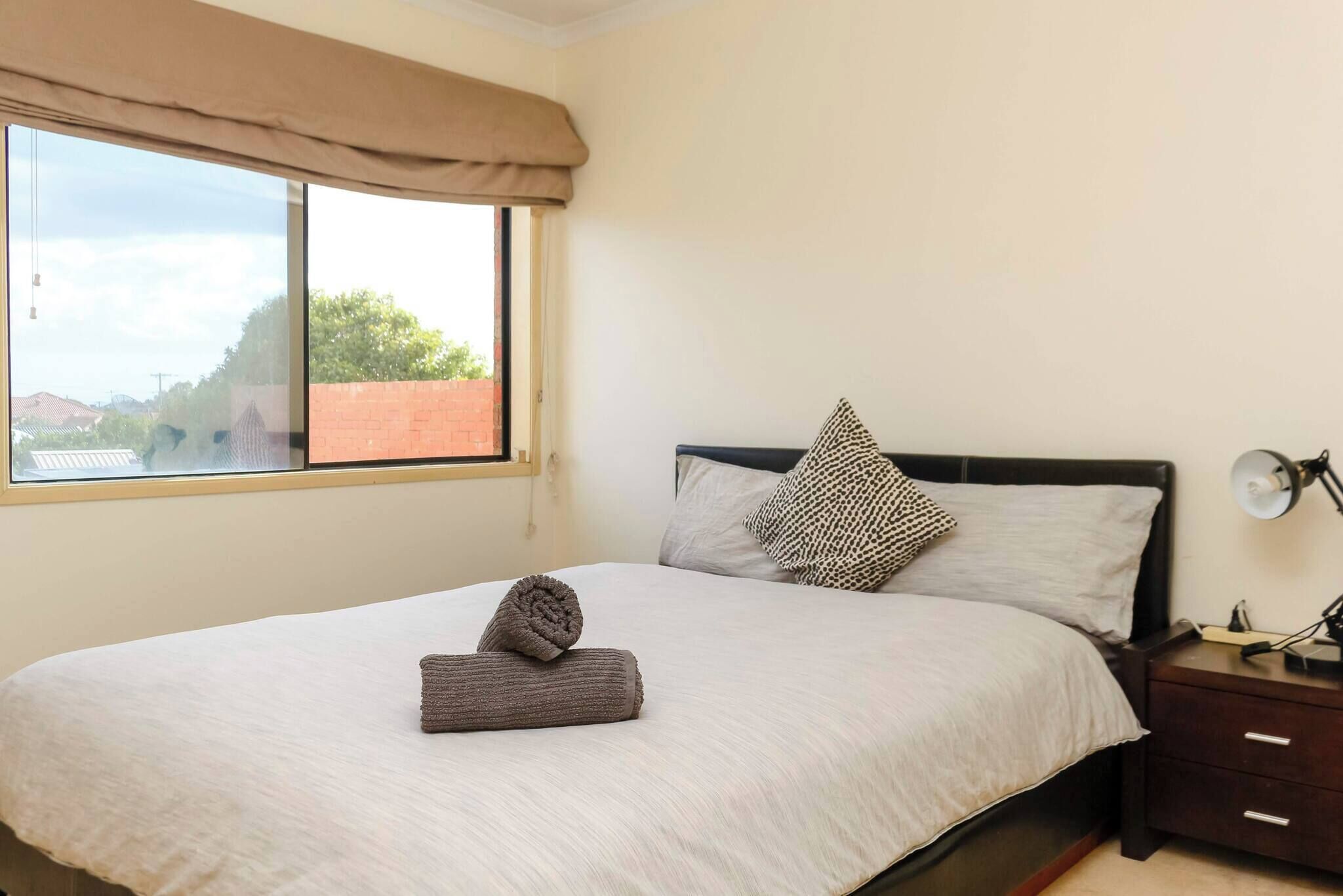 Una cama bien tendida con una toalla enrollada, una ventana con vistas a edificios, una mesita de noche con una lámpara y una pared de ladrillo.