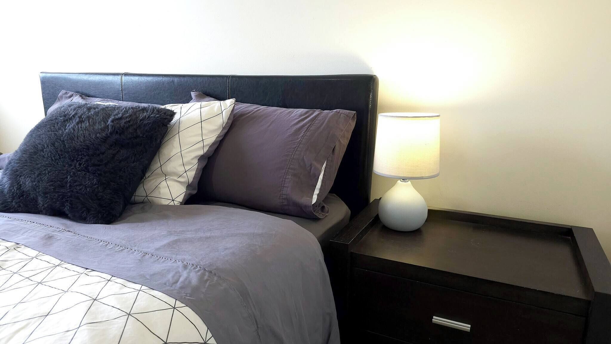 A bed with a geometric-patterned bedspread, a dark headboard, and multiple pillows. There is a white table lamp on a bedside table.