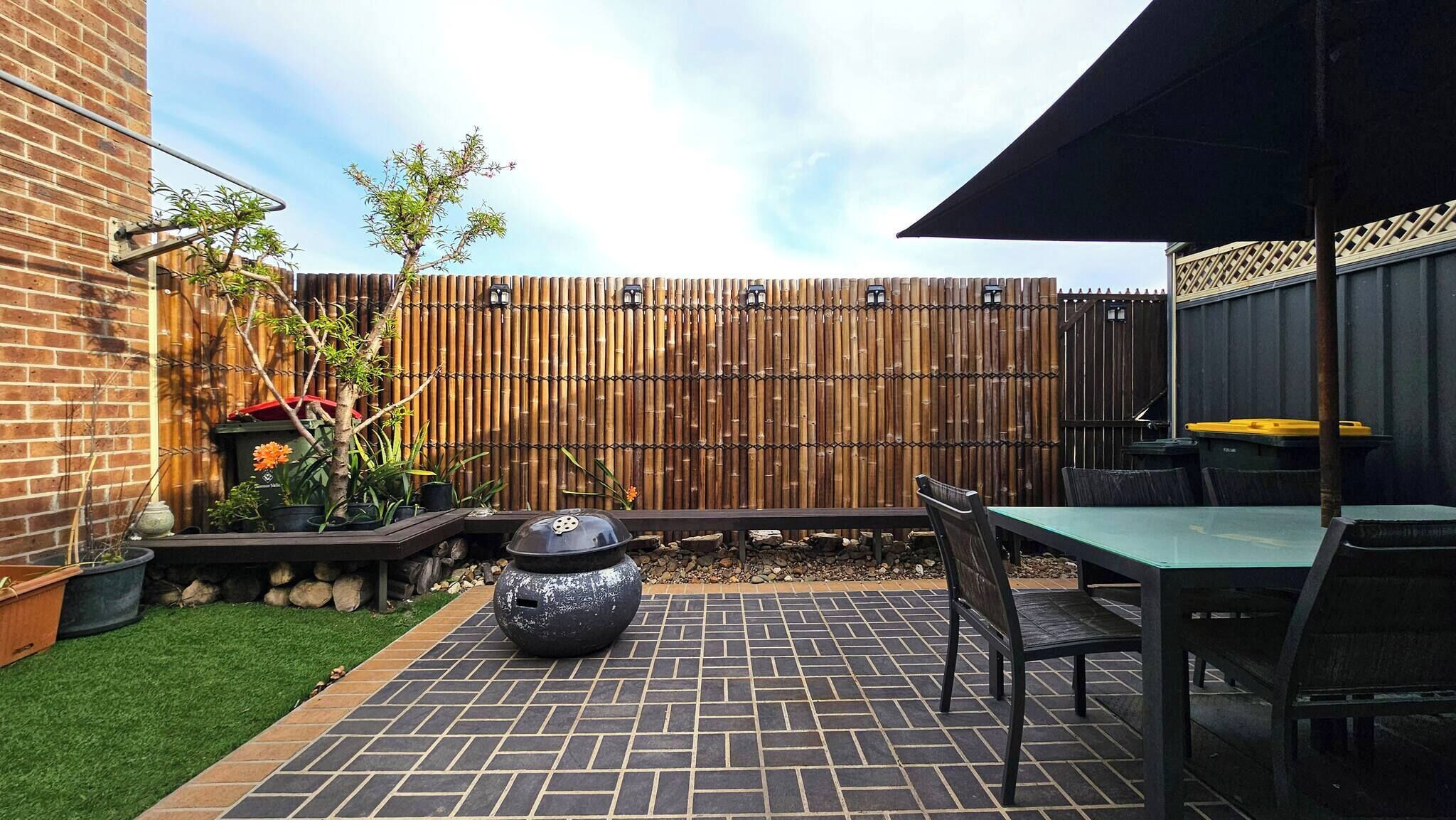 Un patio con suelo de ladrillo, una mesa y sillas, una maceta decorativa y una valla de bambú.
