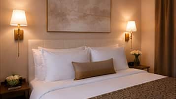 Quarto de hotel com cama, duas luminárias laterais, mesa de cabeceira com um vaso de flores e um quadro na parede.
